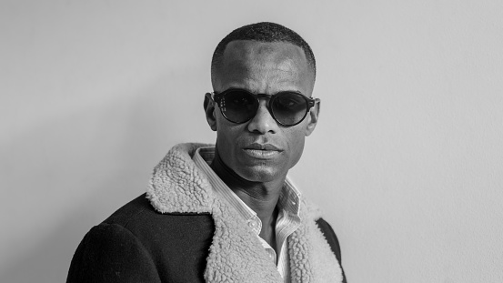 A man stands wearing sunglasses and a blue jacket, looking towards the camera over light solid background high res image