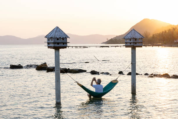Mature man relaxes in hammock suspended over sea as sun rises over distant mountains and uses mobile phone