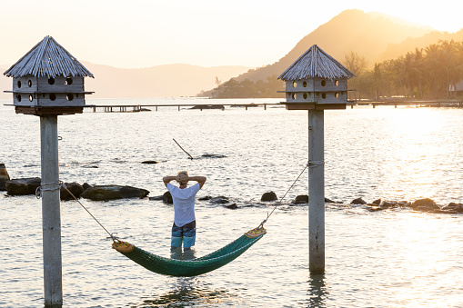 Senior man stands near hammock and watches sun rise over distant mountains from sea