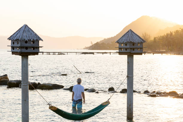 Senior man stands near hammock and watches sun rise over distant mountains from sea