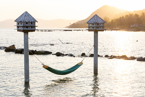 Empty hammock suspended over calm sea as sun rises over distant mountains