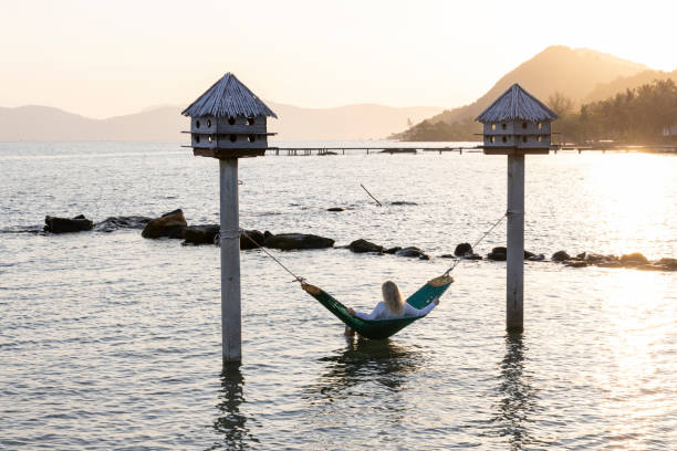 Mature woman relaxes in hammock in morning light suspended over shallow sea water