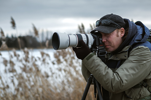 Wildlife photographer is setting up camera on tripod outdoors. Man photographing landscape or animals at lake in winter. Bird watching