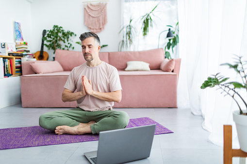 Mid adult man practicing yoga at home online using laptop. Siting in lotus position on exercise mat and meditating with closed eyes