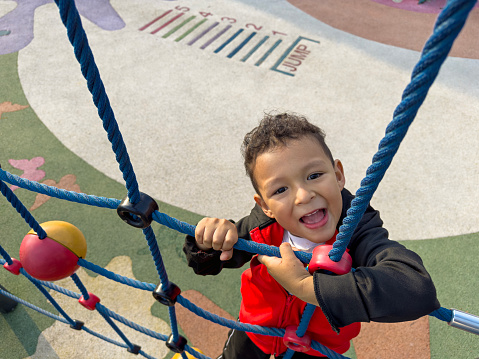 Little boy climbing rope net in an outdoor playground while looking at the camera and smiling, in the background is blue sky on a sunny day.