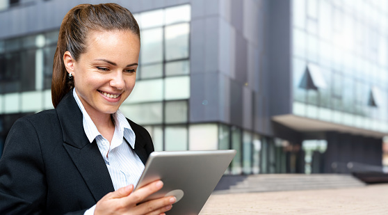 Young brunette businesswoman looking at digital tablet and smiling while standing on the street in front of business building.