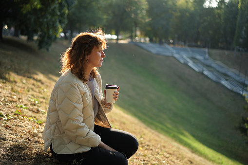 Portrait of young brunette woman with long hair with coffee to go in city park in golden hour. Relaxing, enjoing the sun High quality photo