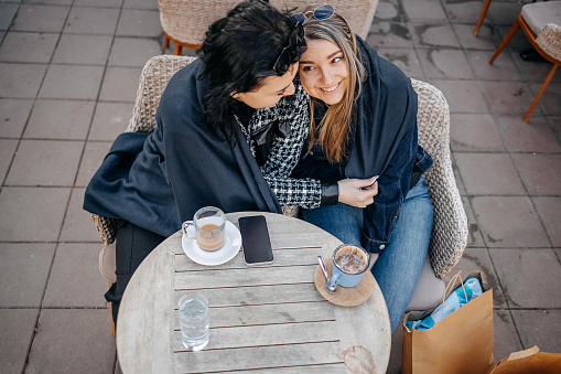 Female friends enjoying while having a coffee break outdoors at restaurant wrapped in a blanket