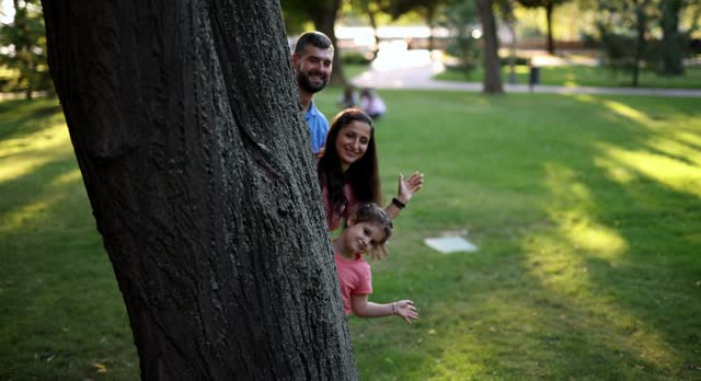 Young Family Standing Behind A Tree In A Sunny Park