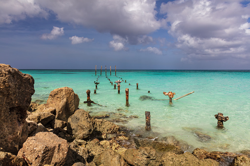 Rocky beach on the island of Aruba. Pylons from an old pier extend to horizon, surrounded by emerald green water. Cloudy blue sky overhead.