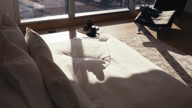 sun shades rising to reveal a bed with white bedding and a bedside tray that includes 2 coffee mugs and a book in a luxury bedroom