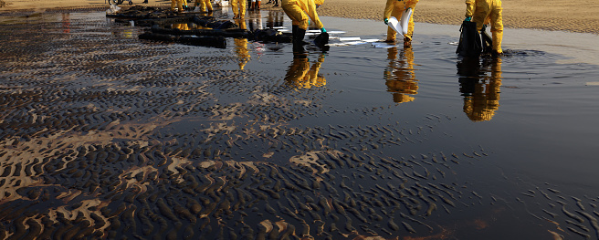 Professional team and volunteer wearing PPE clean up dirty of oil spill on the beach,  oil slick washed up on a sand beach