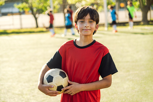 Portrait of a happy young boy holding a soccer ball, with players in the background.