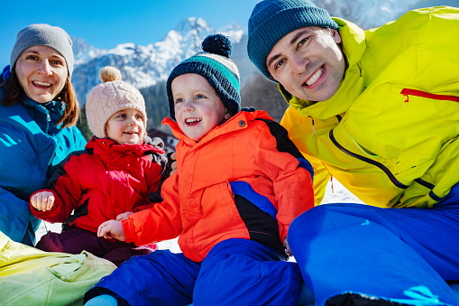 Happy parents with small kids on winter vacations hugging in the snow over peaky mountains laughing looking at camera, cute family portrait