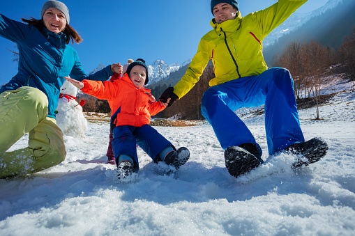 Dynamic shot of two parents jumping in snow with his little son in bright winter outfits over clear sky and mountain chain