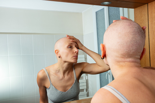 Pensive hairless woman ill of breast cancer in a bathroom, looking at herself in the mirror while touching her bald head