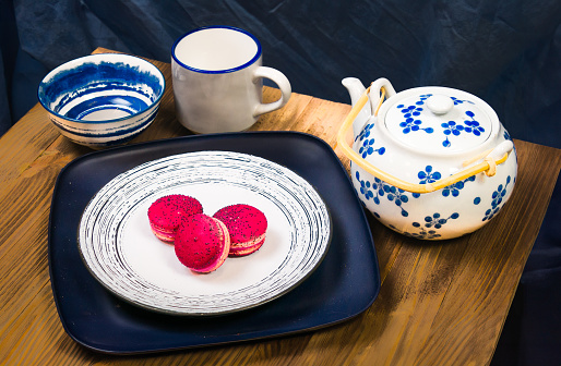 A still life of  three macarons on a plate with a tea pot and cup and finger bowl on a wooden table.