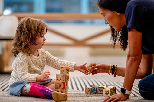 A sweet little girl with a prosthetic leg, sits with her occupational therapist as they play together with wooden blocks.