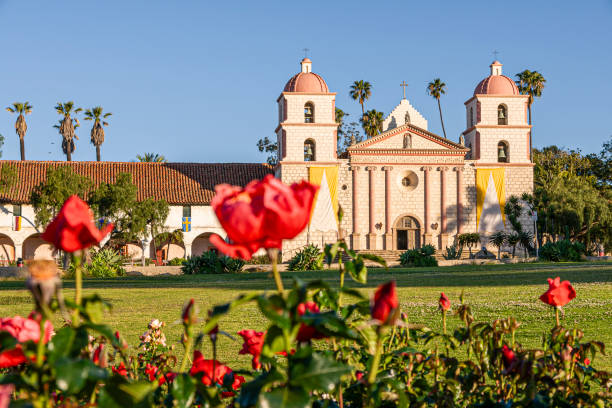 Santa Barbara, CA, USA - April 20, 2009: Old Mission church. from behind red roses under blue cloudscape bathing in sun. Green trees around