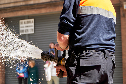 A fireman is seen from behind, handling a fully-engaged hose with a powerful stream of water, likely participating in a training exercise or responding to an emergency.