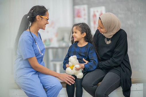 A Muslim Mother sits with her daughter on her lap as they talk with the doctor.  The female doctor of Middle Eastern decent, is dressed professionally in scrubs and smiling as they talk.