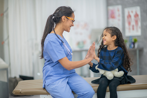 A young girl sits up on an exam table as her female doctor, of Middle Eastern decent, finishes giving her a check-up.  The patient is dressed comfortably and giving the doctor a high-five.