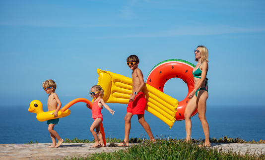 Beautiful family, mother and three kids prepares for a day of swimming, carrying bright inflatables, with a scenic ocean view on the backdrop