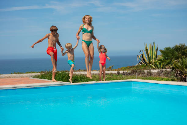 Beautiful family in motion by pool, laugh and jump together An adult and children in bathing suits moments before leaping into a swimming pool, with the sea horizon behind in sunny day person falling backwards stock pictures, royalty-free photos & images