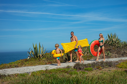 Full height portrait of family, mother, father and two kids equipped with inflatable beach accessories, heading to the oceanfront in bright sunlight