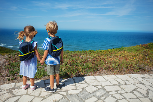 Blond boy and girl with backpacks holding hands admire sea from hill on hot summer day in Portugal