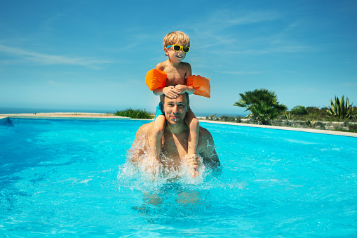 Young child in orange arm floaties and sunglasses rides on the shoulders of an adult male in a sunlit swimming pool