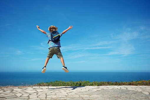 Happy blond kid with backpack and marine t-shirt jump high excited to see ocean on summer hot day