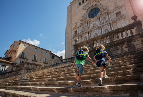 Two cute little tourists with backpacks climb up the ancient stairs of Cathedral of Girona during their summer trip in Spain, view from behind