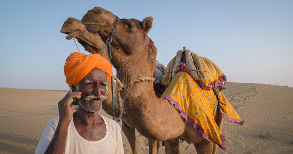 Indian man using a mobile phone on sand dunes with two camels on the background, desert village, Rajasthan, India.