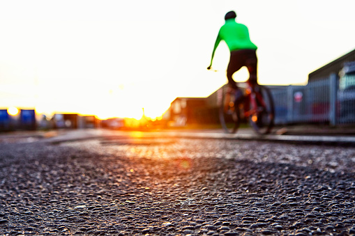 A cyclist, blurred and unidentifiable, rides past a potholed area of road with dawn rising ahead.