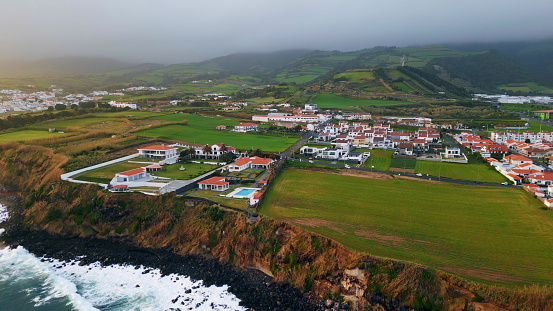 Gloomy coastal resort landscape drone view. Wonderful residential complex with swimming pool placed on picturesque woodland hill. Foaming stormy ocean splashing under rocky shoreline in slow motion