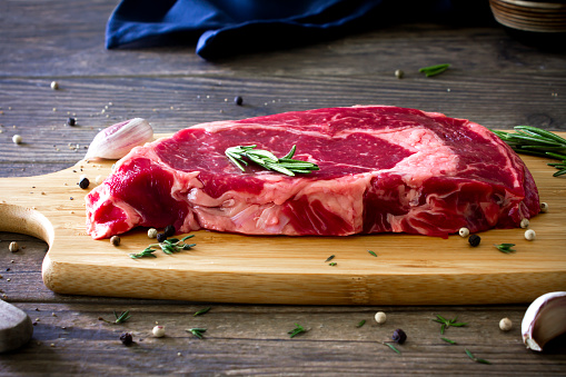 Close up shot of a fresh Ribeye steak on a chopping board, on a rustic wooden kitchen table with rosemary,  garlic and peppercorns.