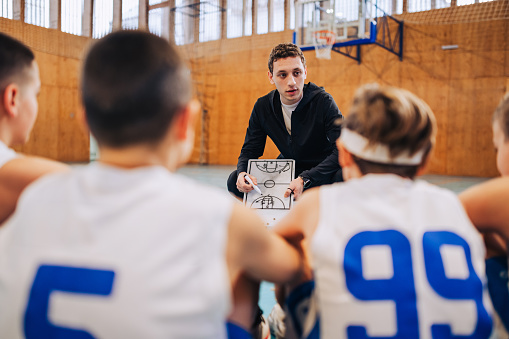 Portrait of a young trainer on basketball court with his junior team and teaching them. A basketball coach with clipboard in his hands is drawing game strategy while teaching his junior basket team.
