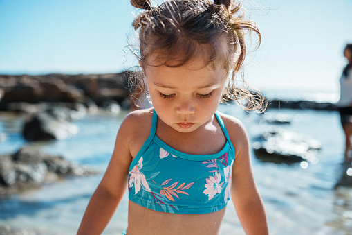 Cropped view of an adorable three year old Eurasian girl looking down with a contemplative expression while playing in at the beach in Hawaii.