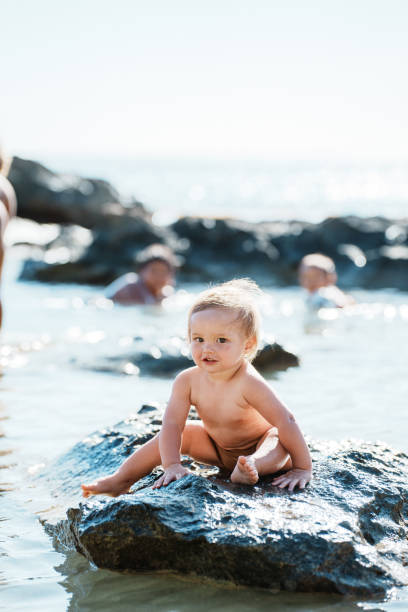 An adorable one year old Eurasian boy of Hawaiian and Chinese descent has fun splashing in the ocean and playing near a rock while at the beach in Hawaii with his family.