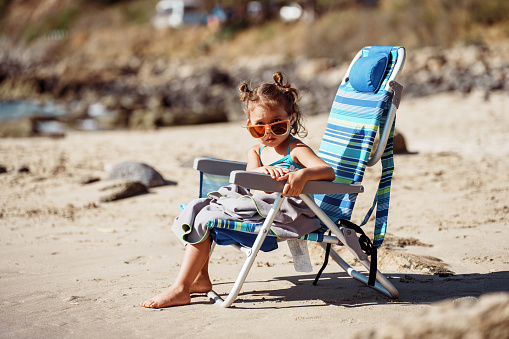 An adorable three year old Eurasian girl wearing a swimsuit and sunglasses relaxes in a folding beach chair on a sandy beach in Hawaii.