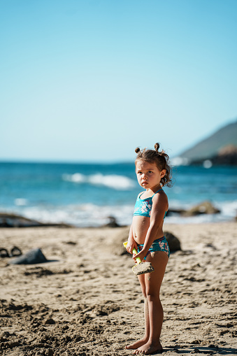 An adorable three year old Eurasian girl of Hawaiian and Chinese descent stand on a sandy beach in Hawaii and holds a shovel while building a sand castle