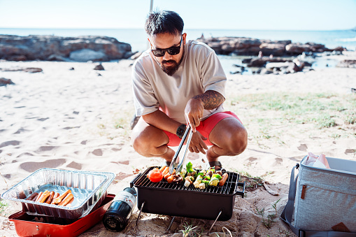 A man of Asian descent squats in the sand at the beach and grills hot dogs and vegetable kabobs on a portable barbecue while enjoying a relaxing day at the beach with family and friends.