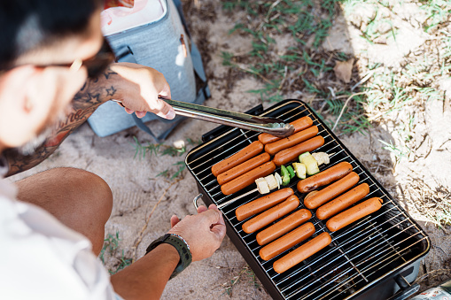 An unrecognizable man uses tongs to cook hotdogs on a portable grill in the sand at the beach in Hawaii while at a barbecue with friends and family.