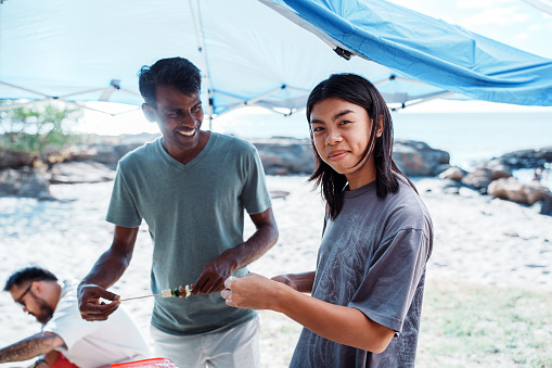 A young man of Asian descent smiles at the camera while grilling with a multiracial group of friends at the beach in Hawaii.