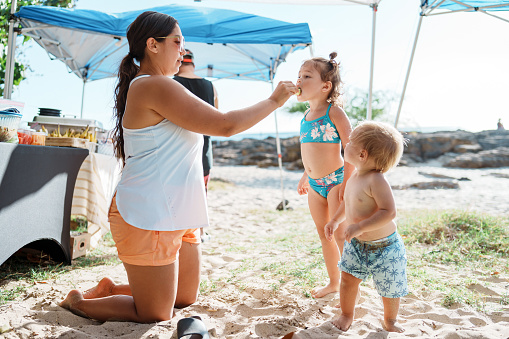 An Hispanic woman feeds her three year old Eurasian niece a bite of food while barbecuing with family and friends at the beach in Hawaii.