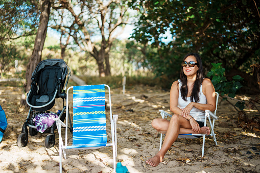 An Asian woman smiles while sitting in a beach chair in the shade of a tree while enjoying a relaxing afternoon at the beach in Hawaii barbecuing with family and friends.