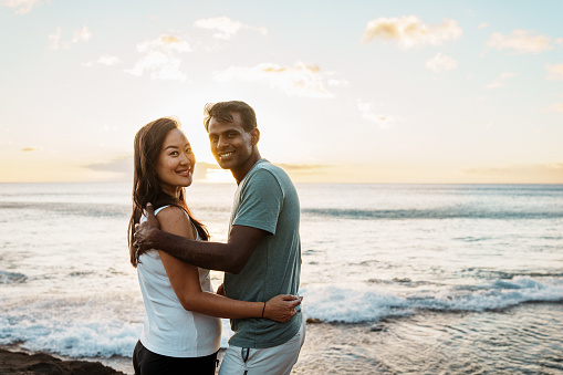 Portrait of an Asian woman and her Indian husband affectionately embracing and smiling directly at the camera while at the beach in Hawaii.