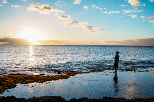 An unrecognizable man stands in the shallow waters of the ocean, holding a fishing pole as he enjoys a sunset on the beaches of Hawaii.