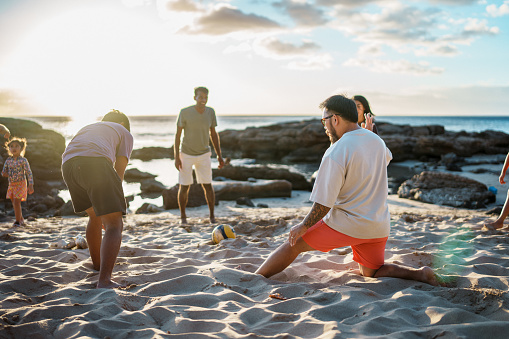 A multiracial group of teenagers and adults play volleyball on a sandy beach in Hawaii during a barbecue with friends and family.